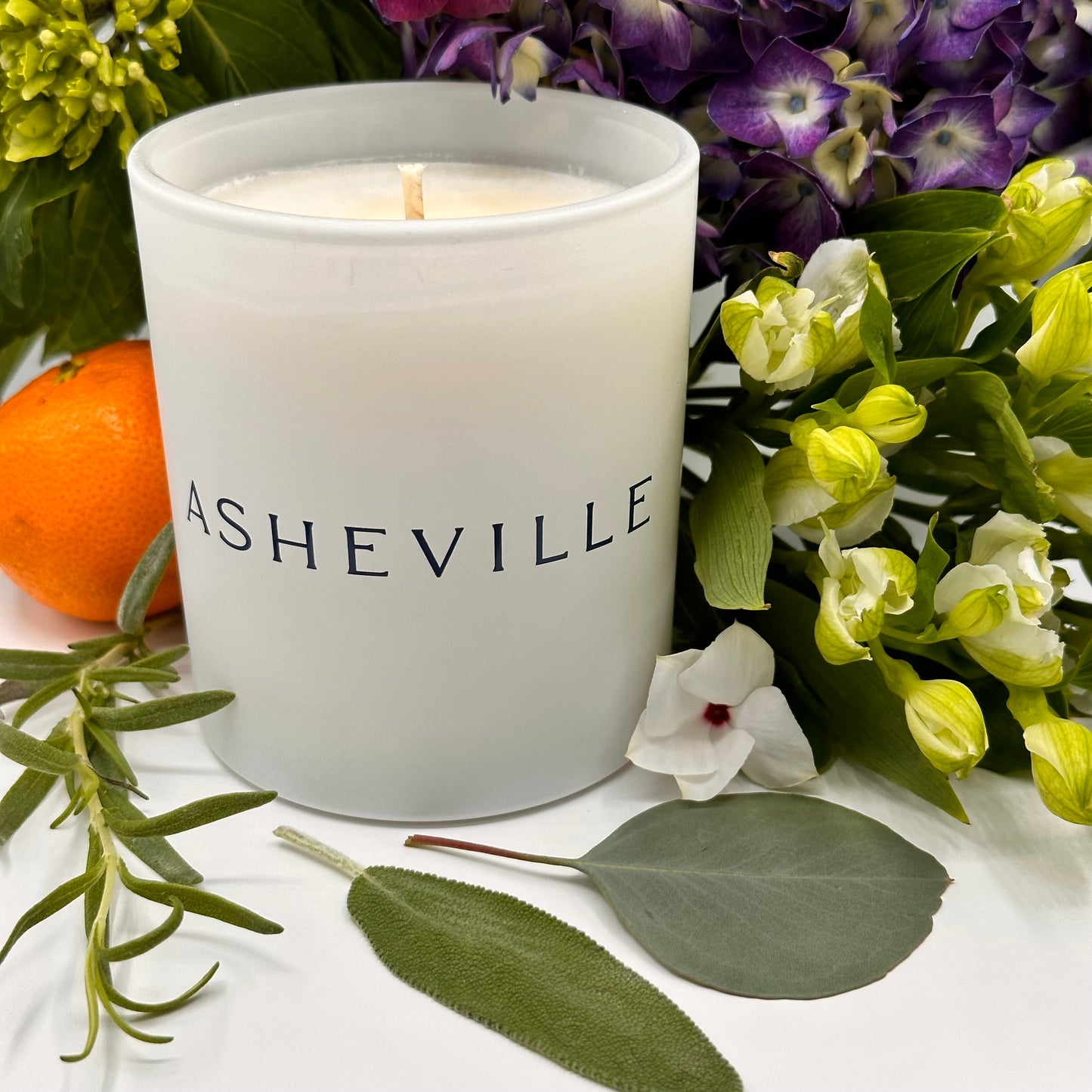 ASHEVILLE Signature Luxury Candle surrounded by flowers orange and herbs